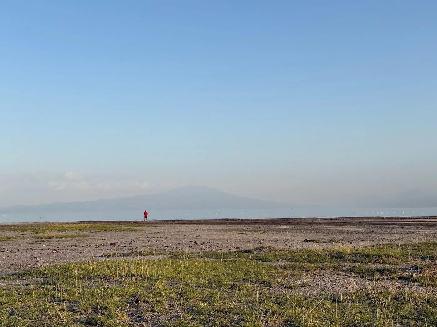 Dry grass and gravel shoreline stretching to a calm lake, with hazy mountains and a small figure on the shore.